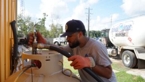 A technician filling a propane tank with a branded truck behind.