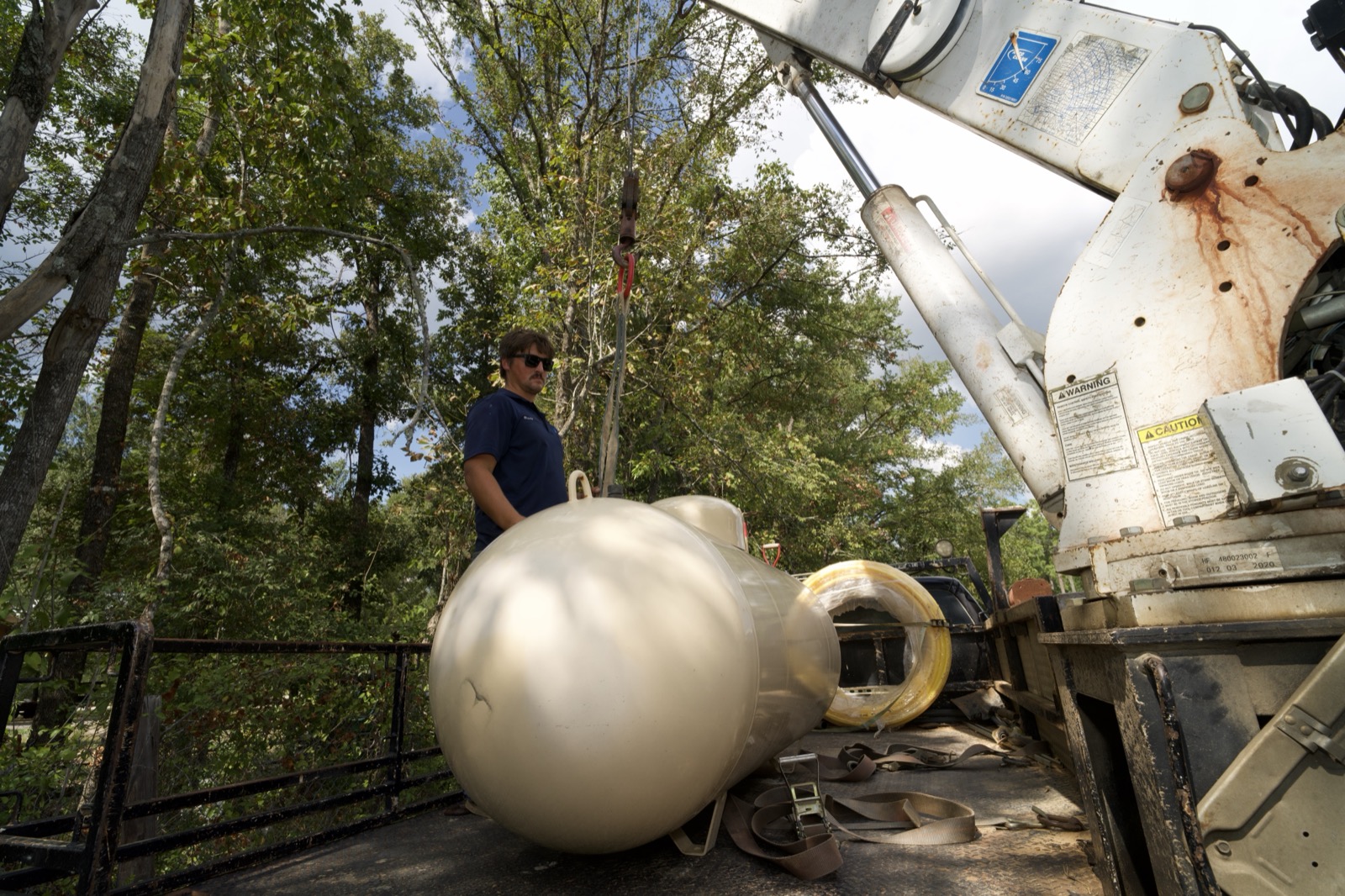 A technician lowering a propane tank from a boom truck at a wooded residential property.