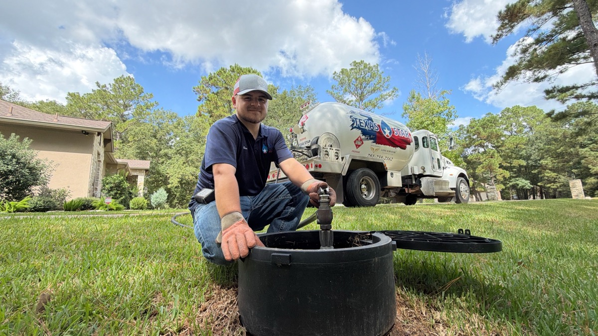 A technician kneels beside an open underground propane tank with a Texas Star Propane bobtail truck parked on the lawn behind him.