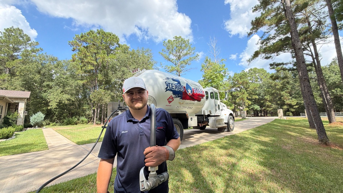 A driver holds a fuel delivery hose nozzle near a Texas Star Propane bobtail truck parked on a tree lined residential driveway.