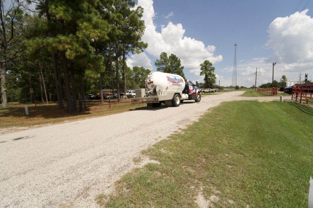 Wide rural shot of propane tanker driving on gravel road; truck is small in frame.